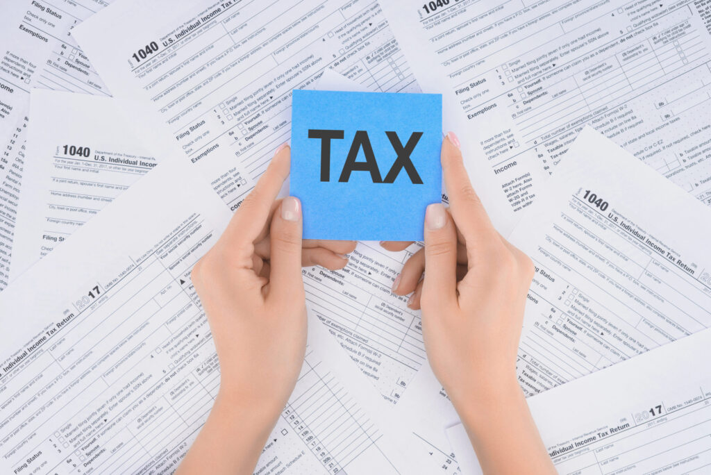 Two hands hold a blue card with "TAX" in bold, surrounded by scattered 1040 tax forms, symbolizing tax filing.